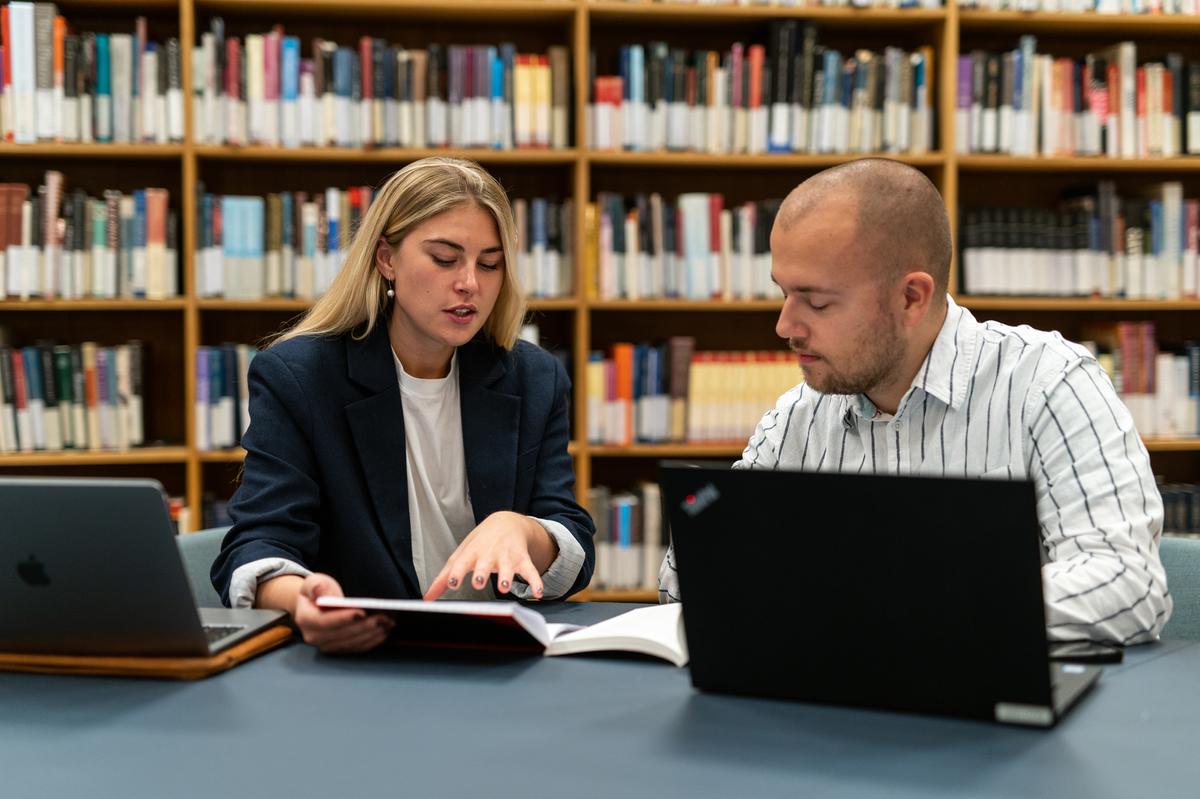 Bibliotek, kvinde og mand sidder med hver sin computer, og kigger i en bog sammen 